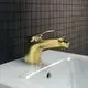 Goldene Bidetarmatur von Ideal Standard mit seitlichem Auslauf und Hebel zum Einstellen des Wasserflusses, montiert auf einer weißen Oberfläche vor dunklem Fliesenhintergrund.